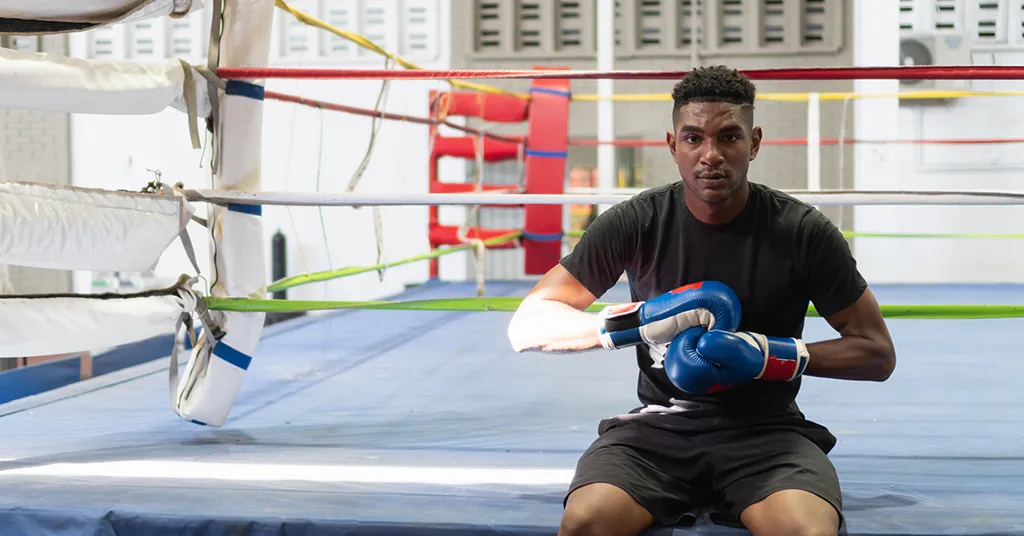 Young male boxer putting on gloves while sitting on boxing ring