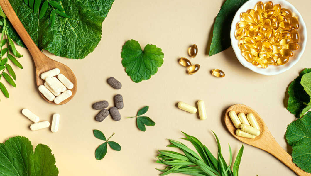 Vegan vitamin capsules for immunity support and healthy lifestyle on light beige background. Natural softgels and pills supplements and various green plant leaves, banner.