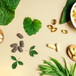 Vegan vitamin capsules for immunity support and healthy lifestyle on light beige background. Natural softgels and pills supplements and various green plant leaves, banner.