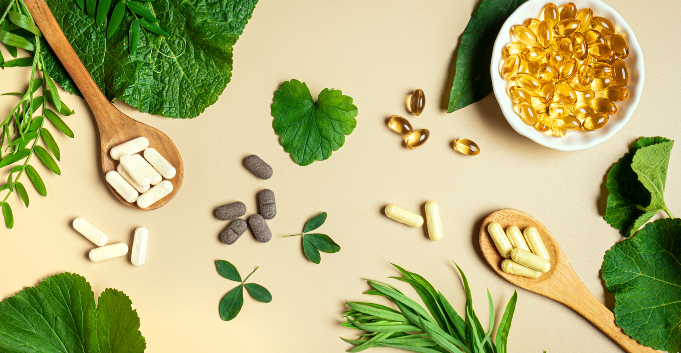 Vegan vitamin capsules for immunity support and healthy lifestyle on light beige background. Natural softgels and pills supplements and various green plant leaves, banner.